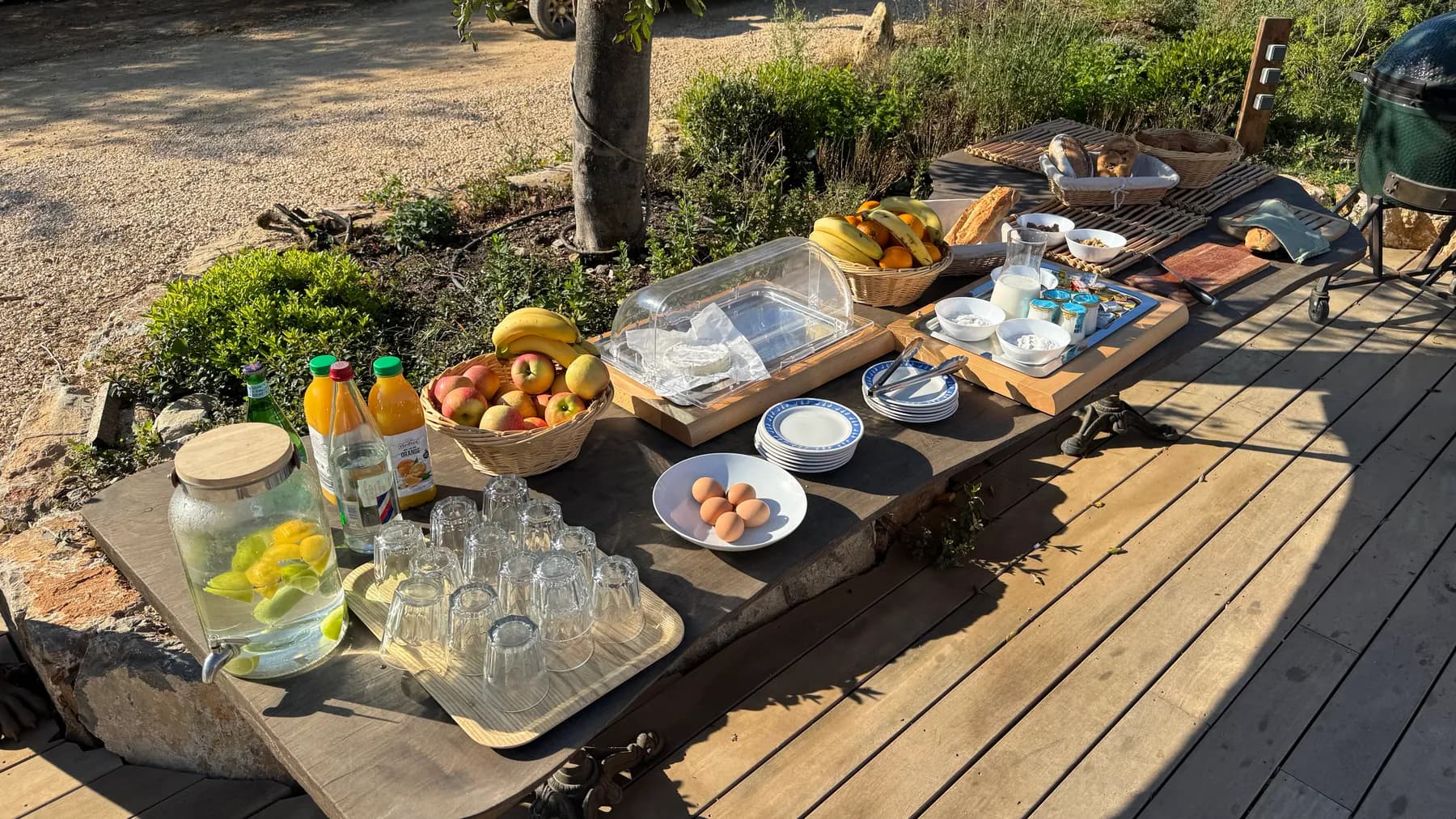 Buffet du petit-déjeuner dressé sur une grande table en bois en terrasse : corbeilles de fruits, œufs, pains et jus d'orange, au soleil du matin